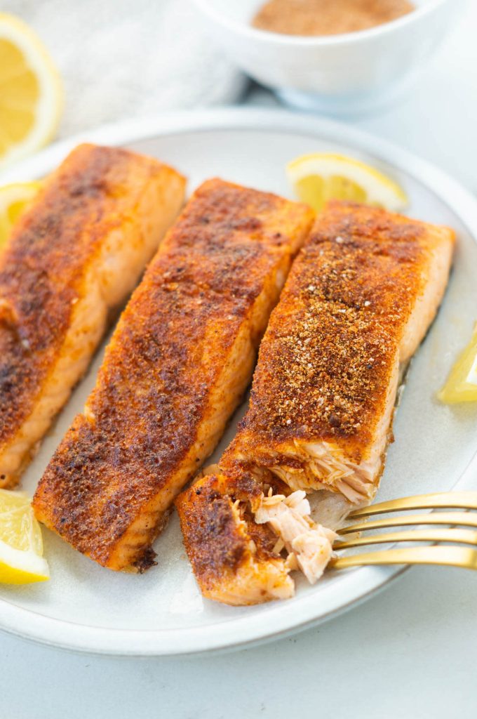 Three filets of blackened salmon lined in a row on a plate. A fork is pulling a piece of the salmon to show its flakiness.