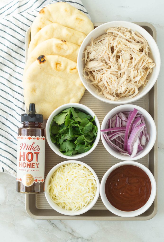 Ingredients needed to make BBQ Chicken Flatbreads. Cilantro, sliced red onion, cheese, shredded chicken and BBQ sauce are in bowls next to four flatbreads and a bottle of Mike's Hot Honey.