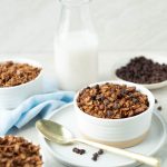 Three bowls of chocolate granola topped with mini chocolate chips and a glass of milk