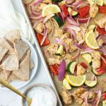 Sheet pan of Greek chicken and veggies topped with lemon slices with a side of pita and yogurt dip