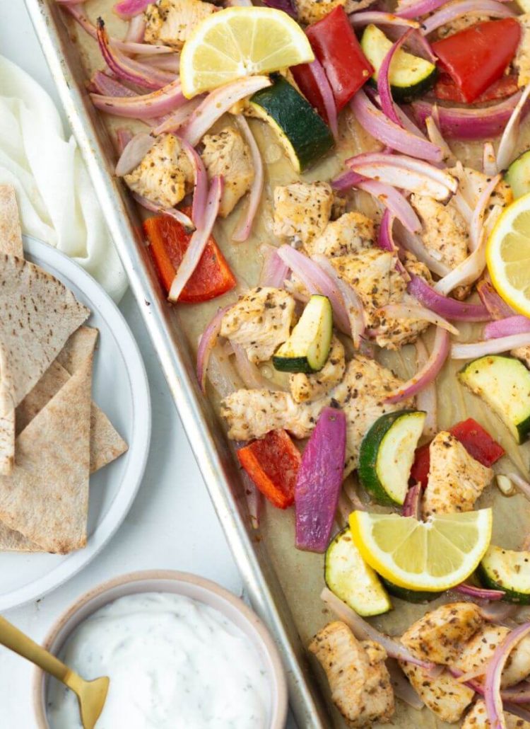 Greek chicken sheet pan with veggies and dill sauce and pita chips on the side.