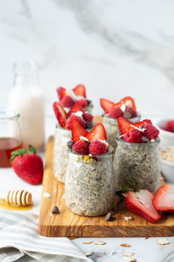 Jars of protein overnight oats topped with berries, chocolate chips, and a drizzle of honey sitting on a wooden board.