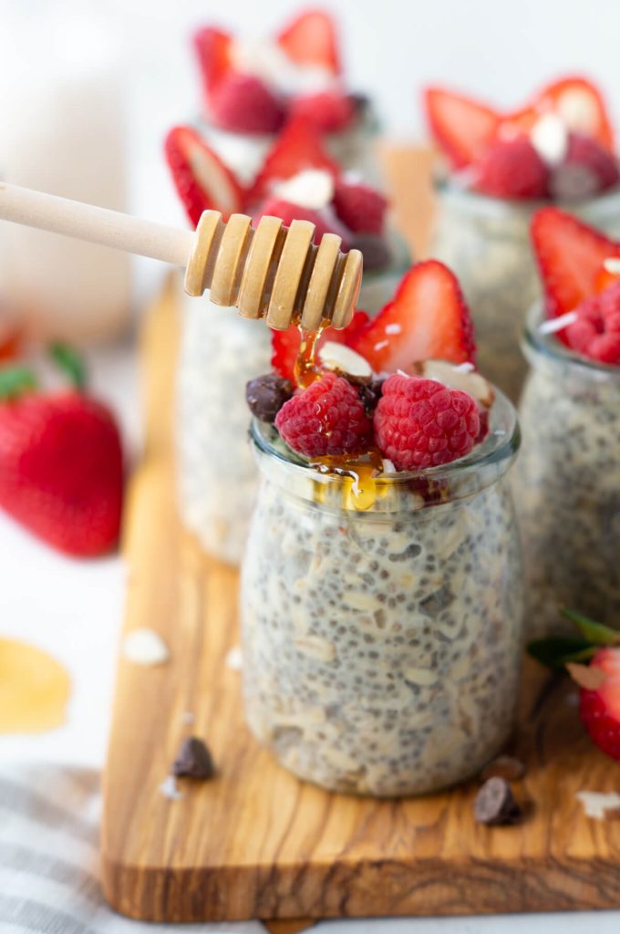 Jars of protein overnight oats topped with berries, chocolate chips, with honey being drizzled on top.