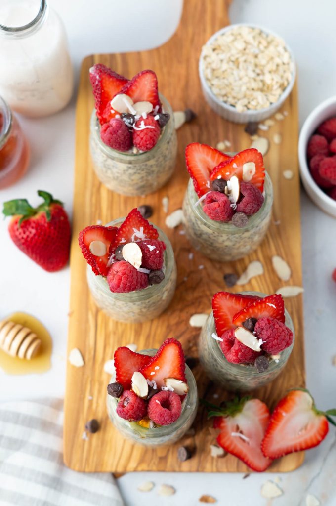 Overhead shot of five jars of protein overnight oats topped with berries, chocolate chips, and a drizzle of honey.