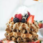 Syrup being poured on a stack of banana protein waffles with berries and whipped cream