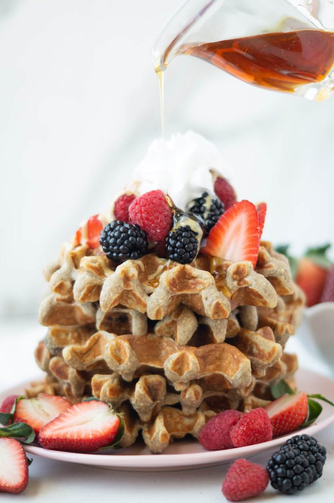 Syrup being poured on a stack of banana protein waffles with berries and whipped cream