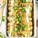 Overhead shot of a pan of sweet pork enchiladas topped with diced tomatoes, red onion, avocado, sliced jalapeños, and sour cream.