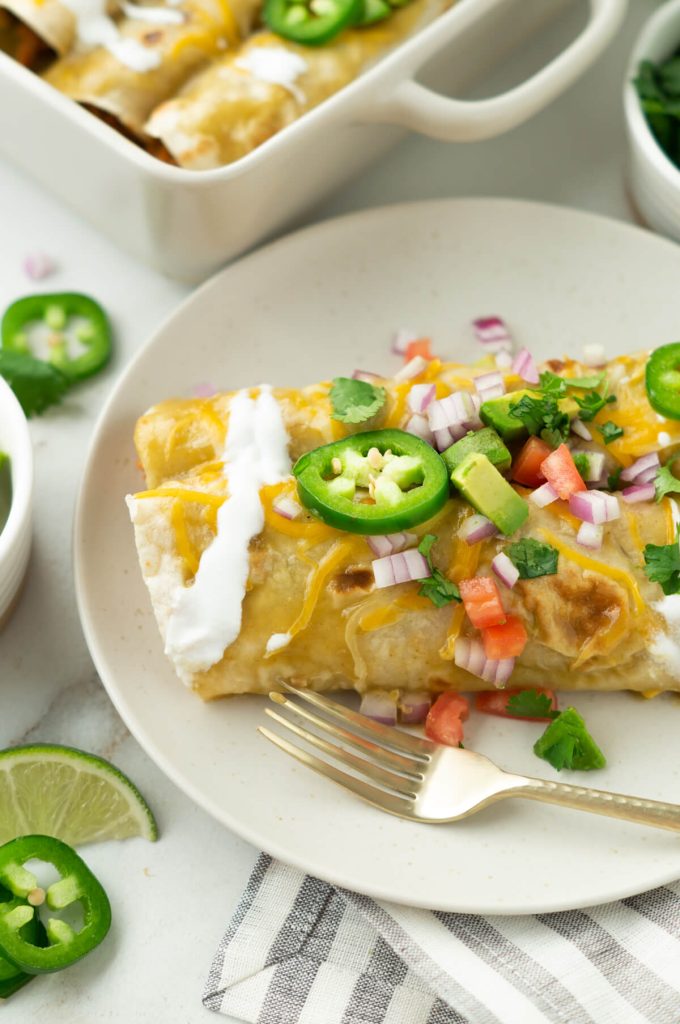 A plate of two sweet pork enchiladas topped with diced tomatoes, red onion, avocado, sliced jalapeños, and sour cream.