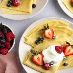 Overhead photo of three plates with crepes, whipped cream, berries, and a bowl of berries and a pink napkin on the side.
