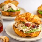Shot of a croissant filled with Chicken salad sandwich and lettuce with honey, a pink napkin, and cashews to the side.