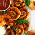 An overhead shot of a platter stacked with homemade air fryer pizza rolls with a bowl of marinara sauce, parmesan cheese, and a garnish of basil leaves.