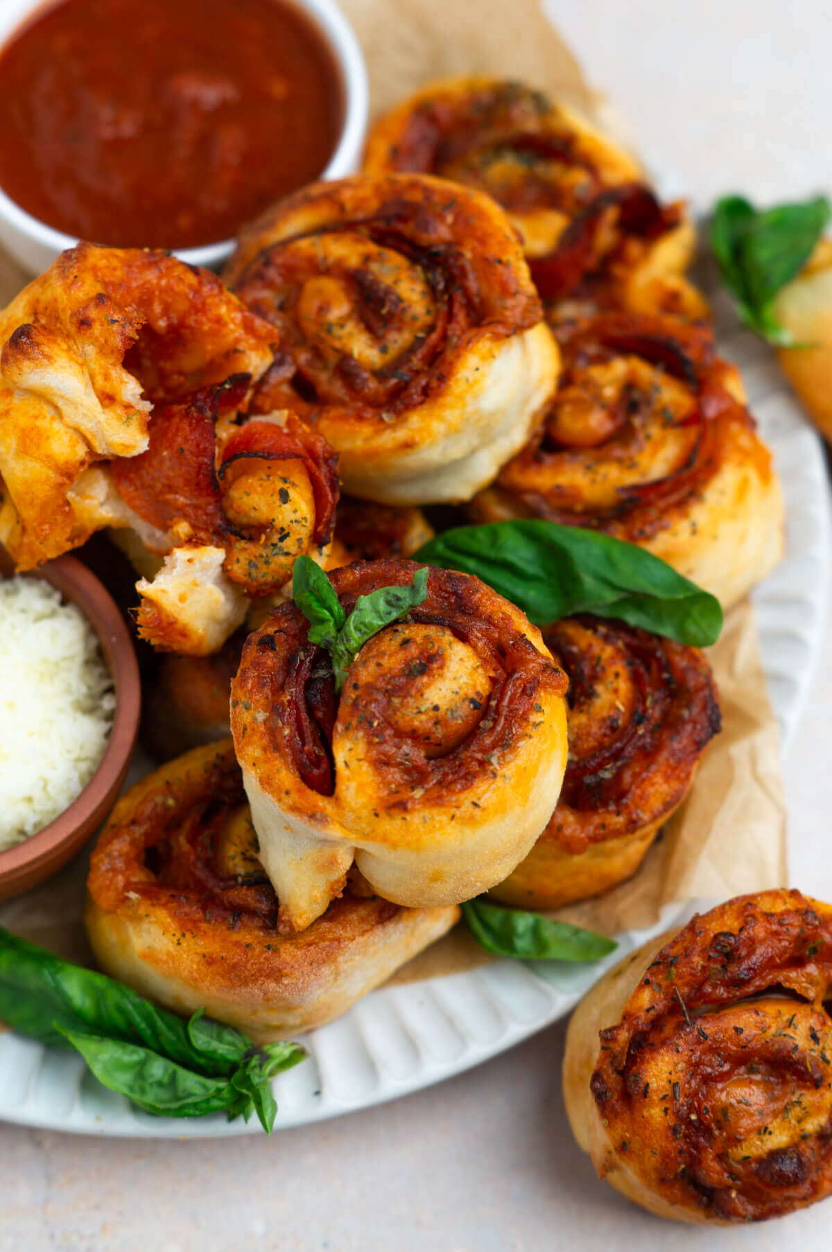 A platter stacked with homemade air fryer pizza rolls with a bowl of marinara sauce, parmesan cheese, and a garnish of basil leaves.