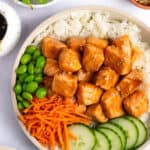 A bowl with white rice, teriyaki salmon bites, edamame, shredded carrots, and sliced cucumber.