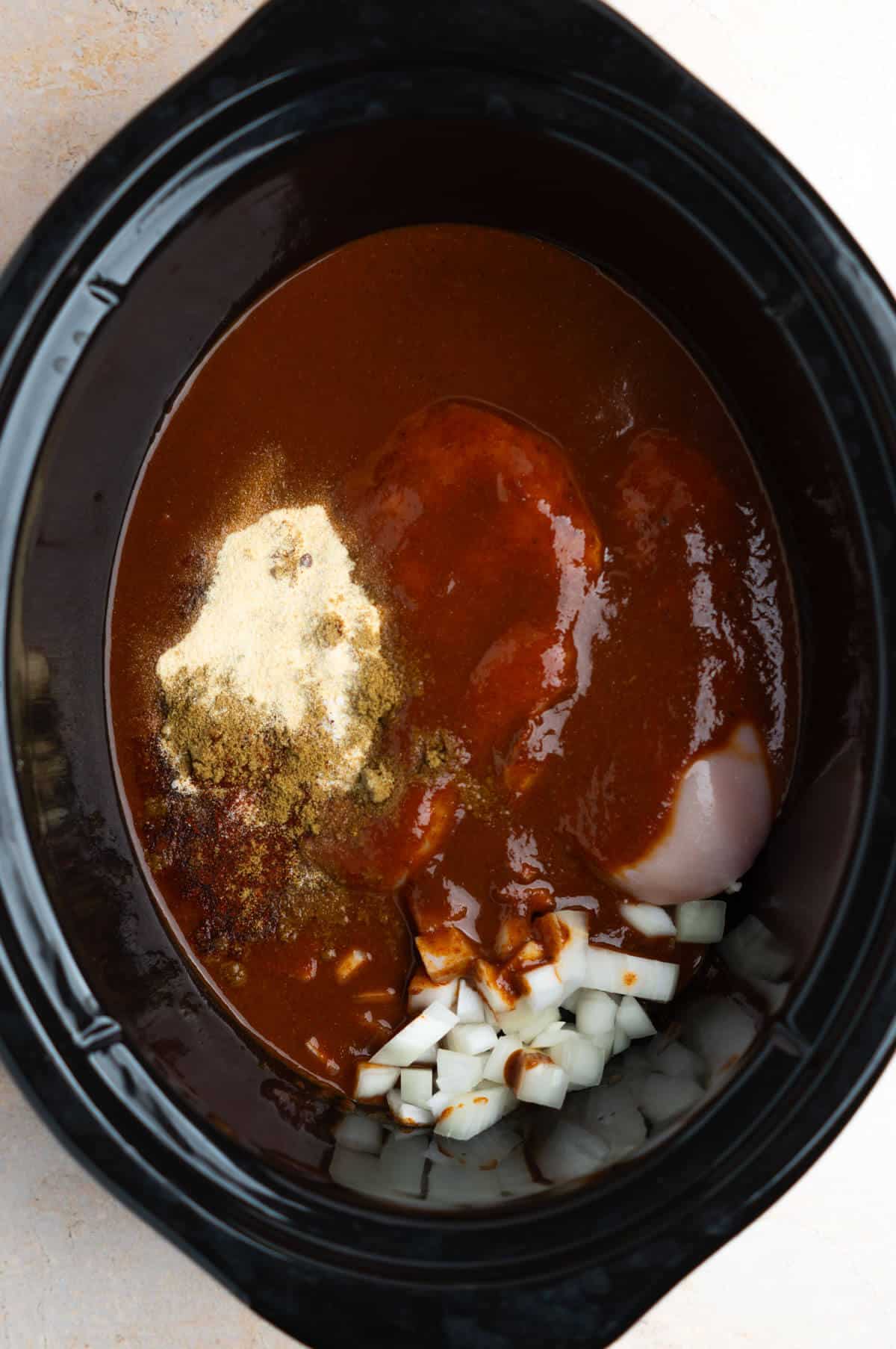 An overhead shot of the slow cooker with the chicken, spices, onion, and red enchilada sauce inside.