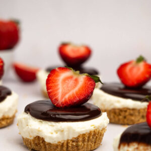 A scattered row of no bake mini cheesecakes topped with chocolate ganache and a strawberry half.