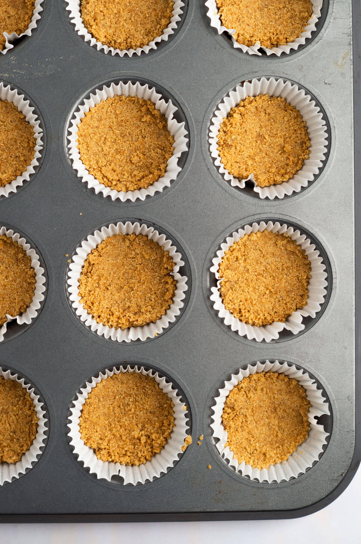 An overhead shot of a muffin tin lined with muffin liners and filled with graham cracker crust.