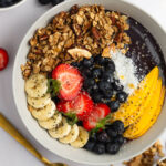 Overhead shot of a protein acai bowl topped with granola, banana, strawberries, blueberries, mangoes, and coconut flakes.