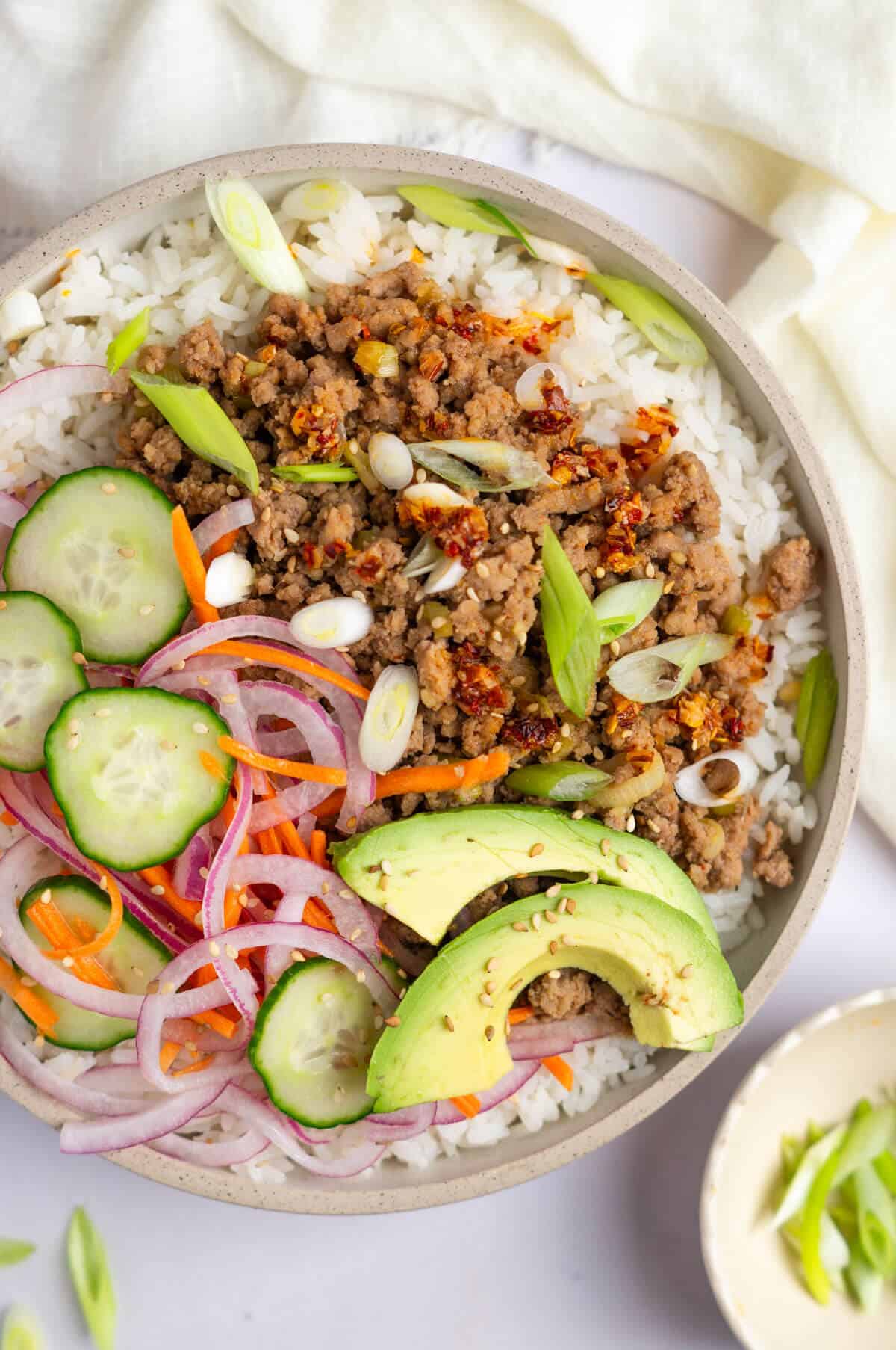 An overhead shot of a bowl with white rice, Korean ground turkey, pickled veggies, avocado slices, and sliced shallots.