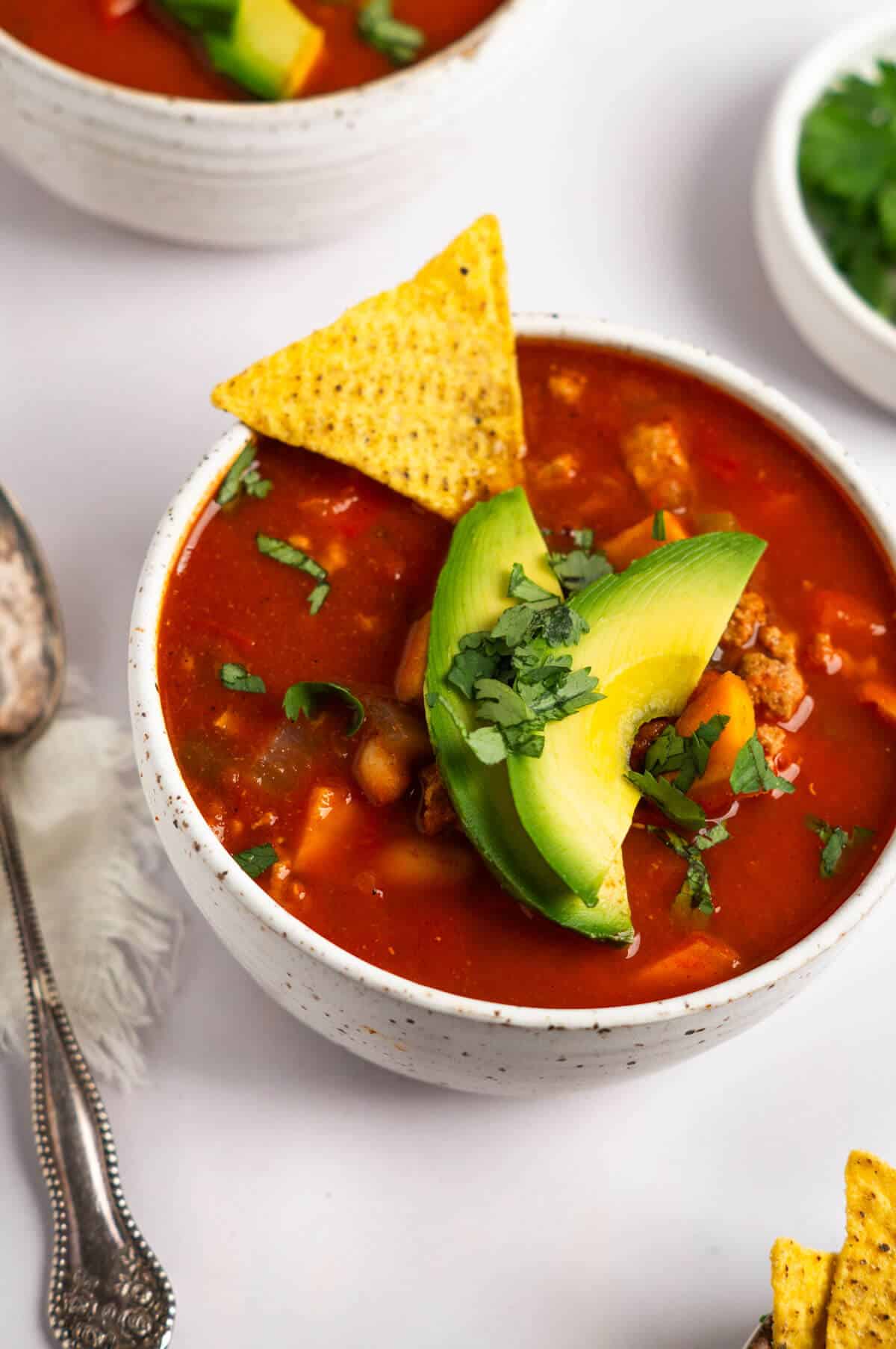 A bowl of turkey sweet potato chili garnished with avocado slices, chopped cilantro, and tortilla chips.