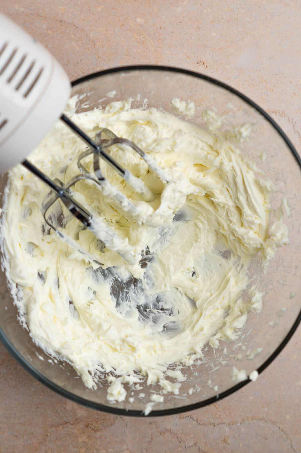 A large mixing bowl with cream cheese that is mixed by an electric hand mixer.