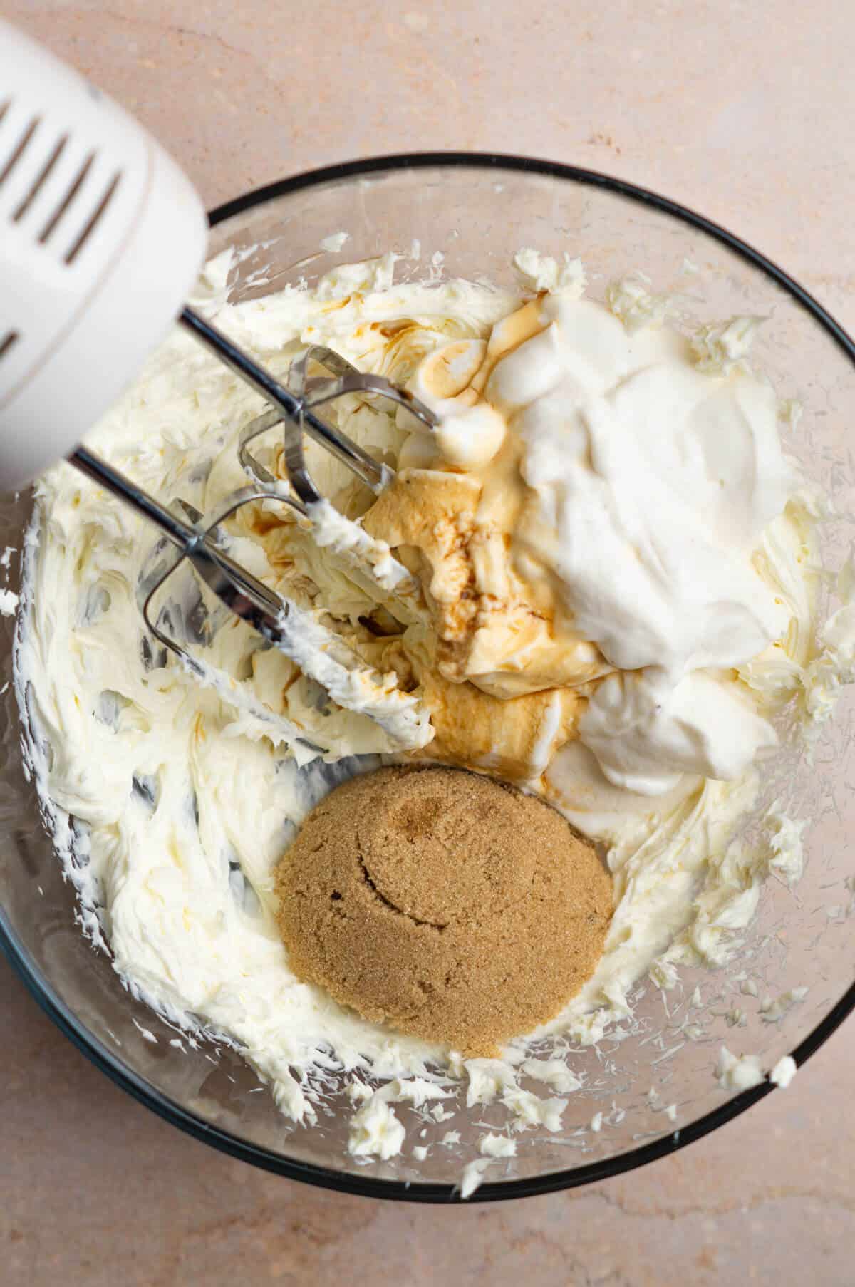 A large mixing bowl with cream cheese, vanilla Greek yogurt, brown sugar, vanilla extract, and a pinch of salt that is being mixed together with an electric hand mixer.