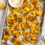 A parchment-lined baking sheet with garlic parmesan smashed potatoes garnished with chives and a small bowl of freshly grated parmesan.