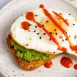 A plate with hash brown avocado toast topped with hot sauce and everything but the bagel seasoning.