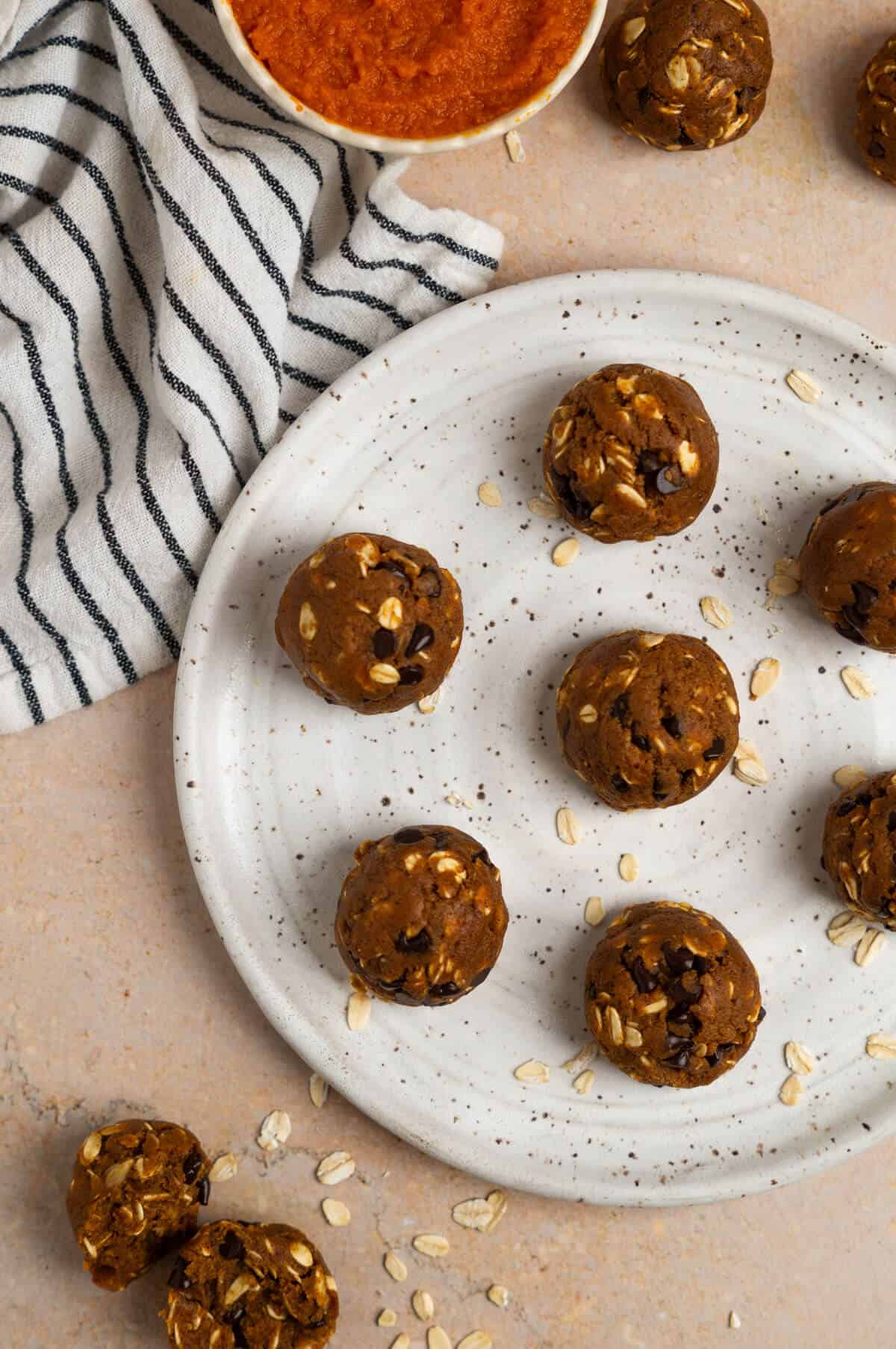 An overhead shot of a plate with seven pumpkin protein balls with a bowl of pumpkin puree and a striped linen cloth in the background.