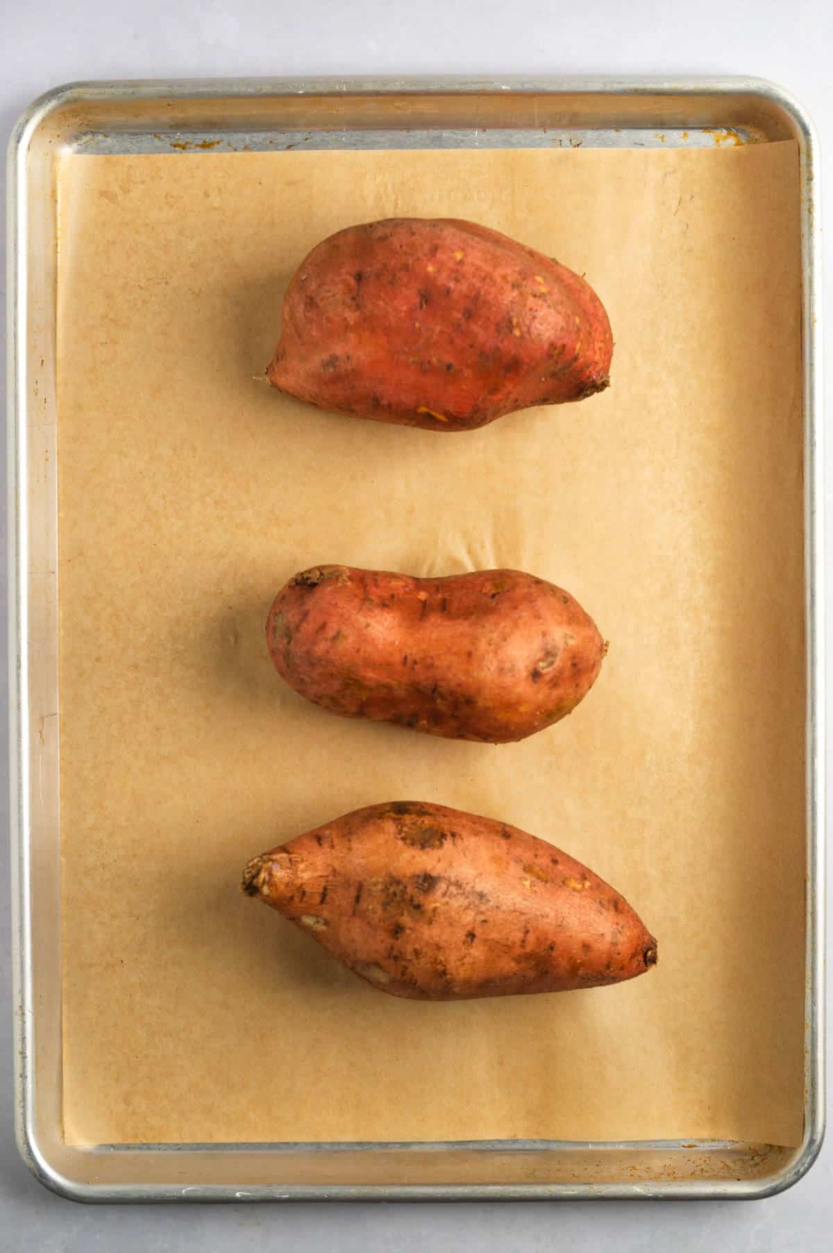 Three sweet potatoes in a line on a parchment-lined baking sheet.