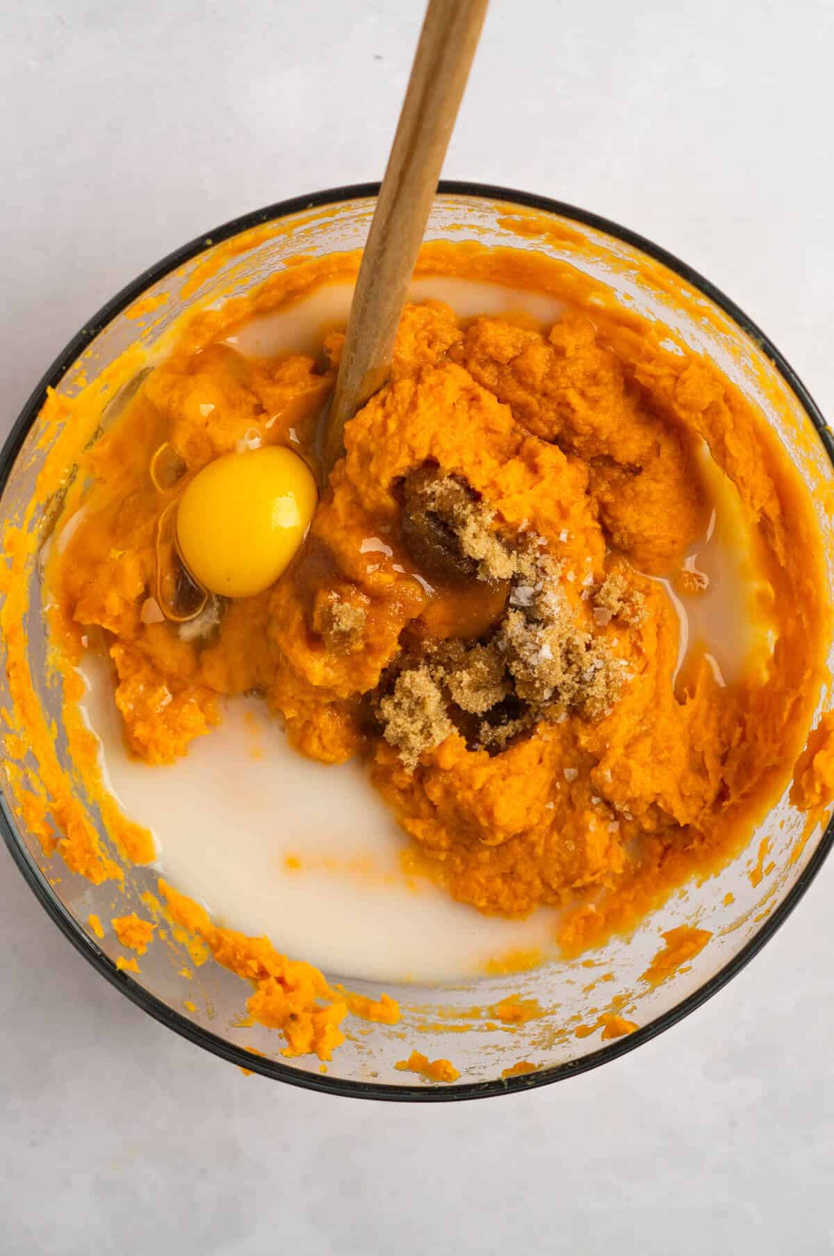 A mixing bowl with mashed sweet potatoes, an egg, milk, brown sugar, cinnamon, vanilla extract, and salt.