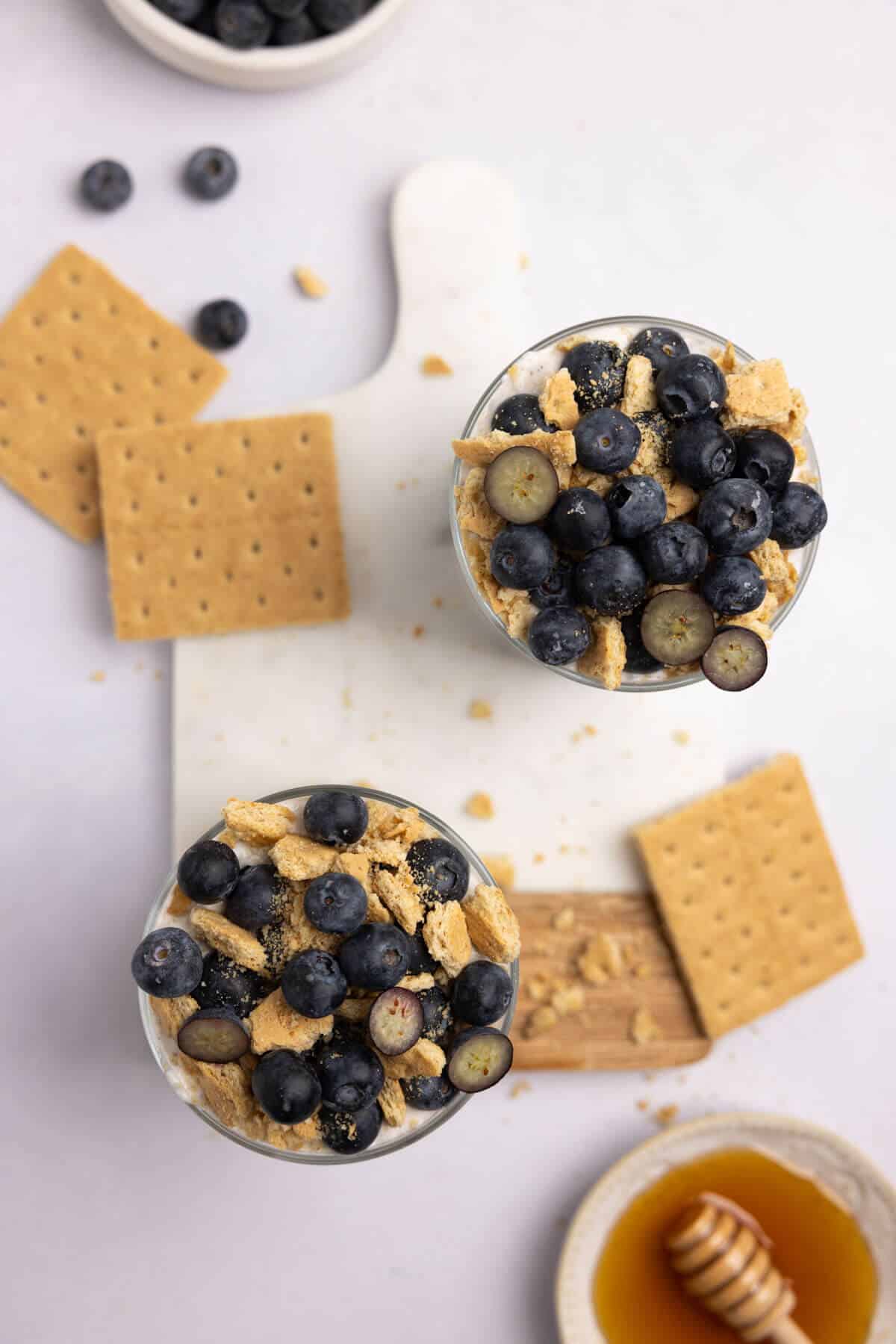 An overhead shot of two cups of overnight oats with blueberries and graham cracker crumbs on top.