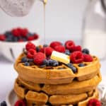 A stack of waffles with a pat of butter, berries, and maple syrup being drizzled over the top.