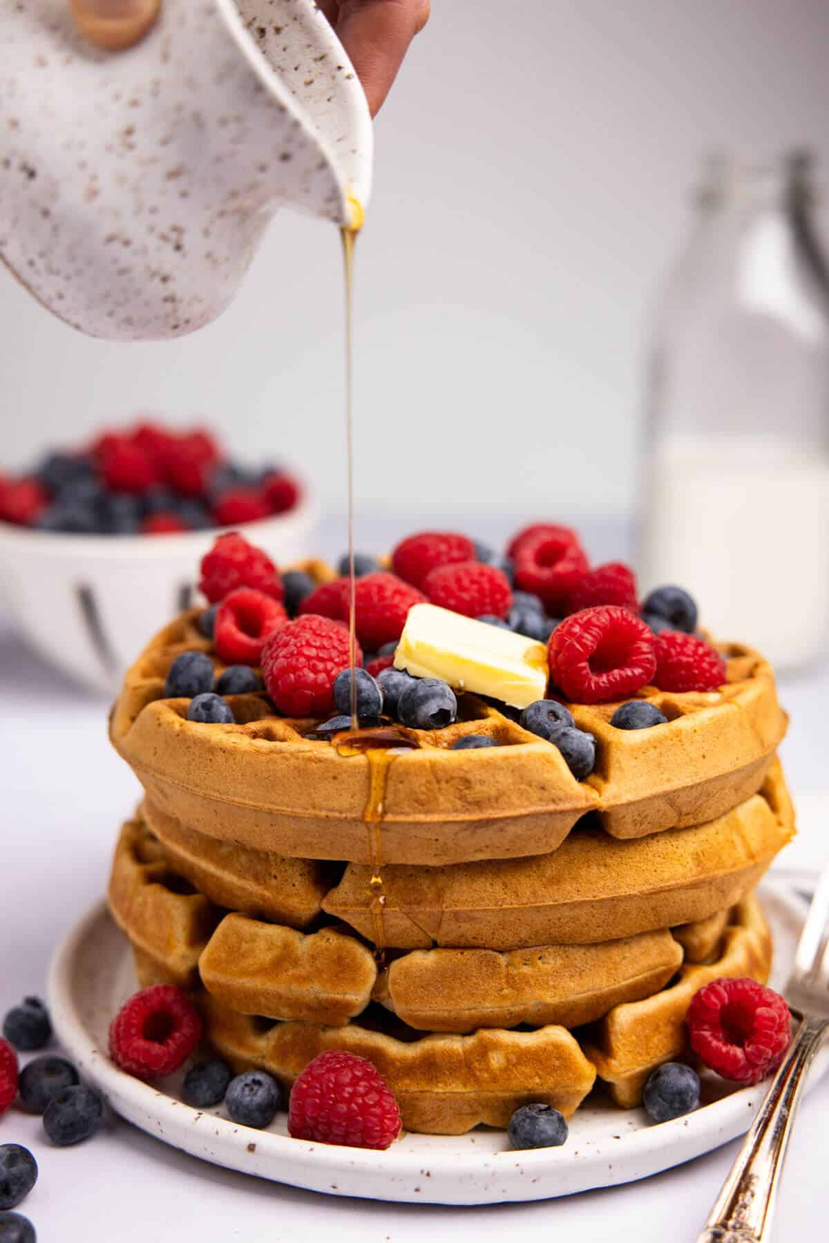 A stack of waffles with a pat of butter, berries, and maple syrup being drizzled over the top.