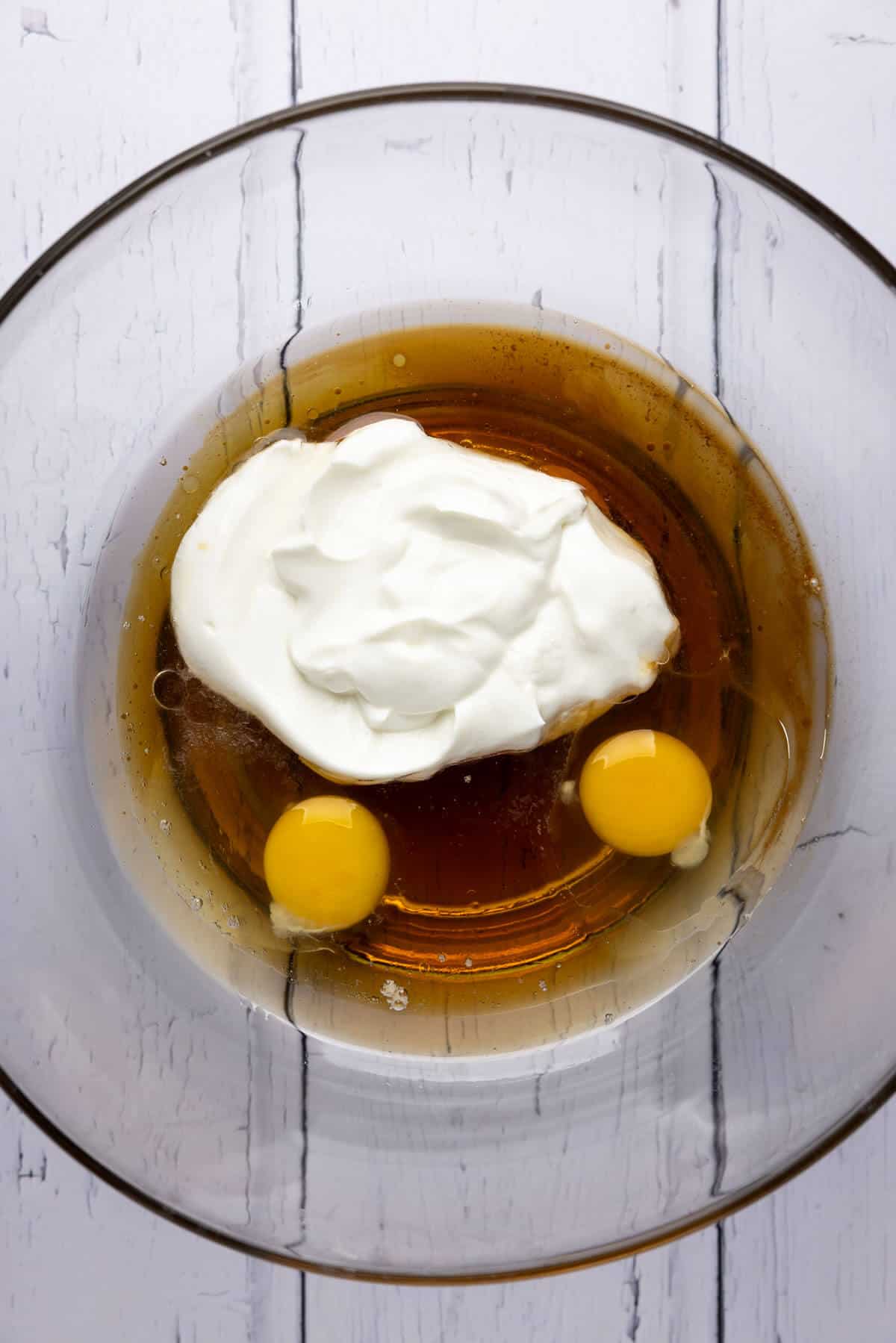 A large mixing bowl with Greek yogurt, eggs, maple syrup, oil, and vanilla extract.