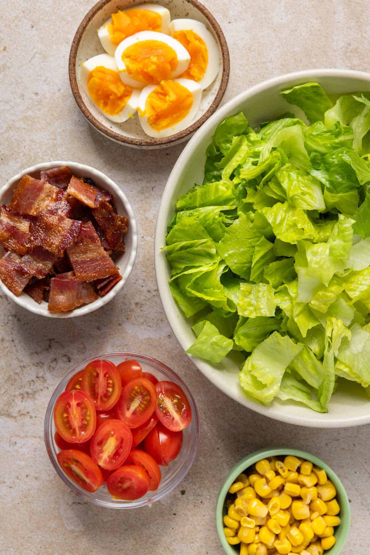 A large bowl of romaine lettuce and small bowls of hard boiled eggs, bacon pieces, grape tomatoes, and corn.