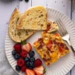 A plate with a piece of egg casserole, fresh fruit, and a slice of sourdough bread.
