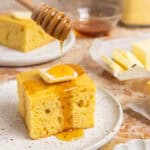 A plate with a piece of cornbread with butter and honey drizzled on top.