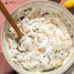 A large mixing bowl of whipped cream fruit salad with a wooden spoon.
