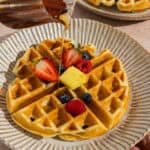 A plate with a cottage cheese waffle topped with berries and a pat of butter with maple syrup being drizzled over the top.