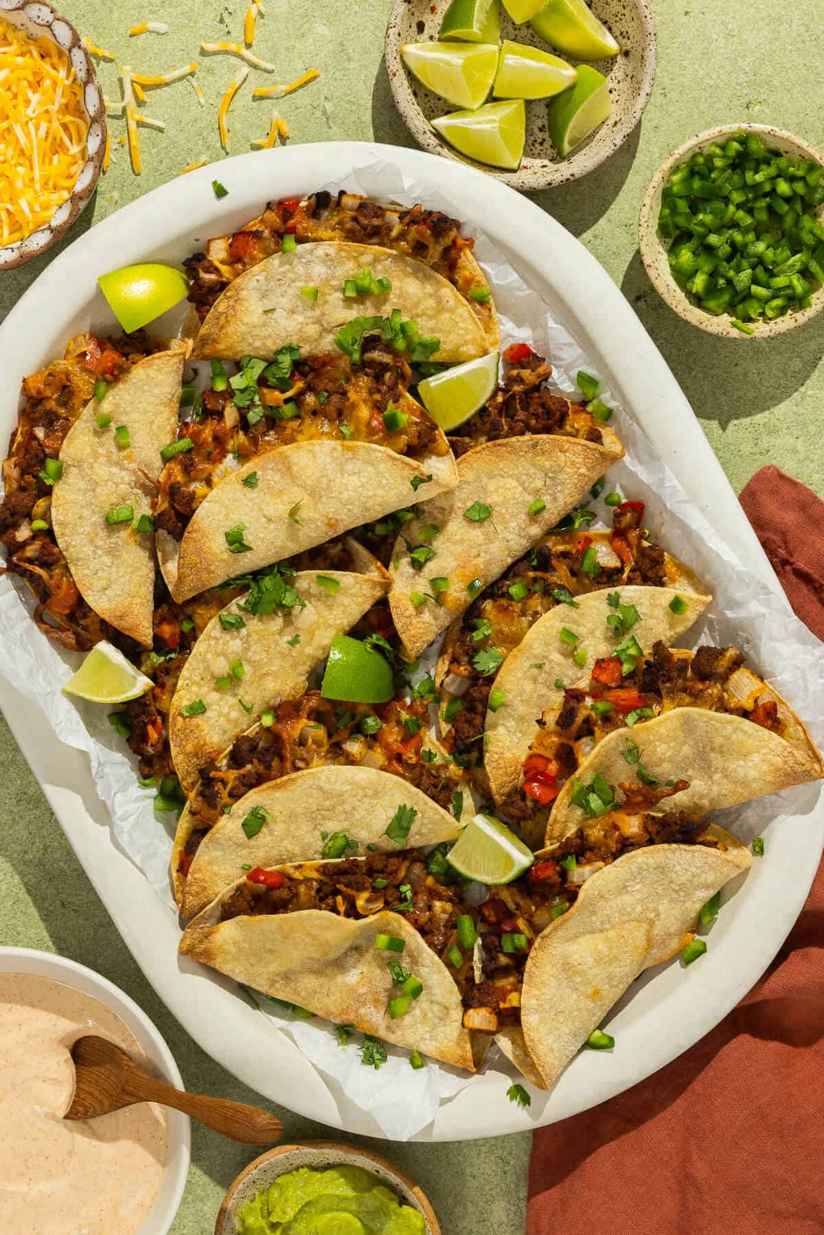 A platter of baked beef tacos garnished with diced jalapeรฑo, cilantro, and lime wedges.