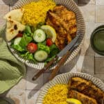 Two plates with Greek lemon chicken, lemon rice, a Greek salad, and pita bread.