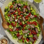 A large platter of tri tip steak salad and serving spoons.