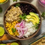 A bowl with salsa verde chicken, rice, beans, corn, avocado, and red onion.