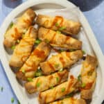 A platter with baked buffalo chicken taquitos garnished with buffalo sauce and sliced green onions.