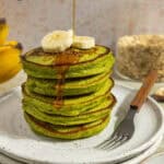 A plate with a stack of banana spinach pancakes topped with bananas and with maple syrup being drizzled over the top.