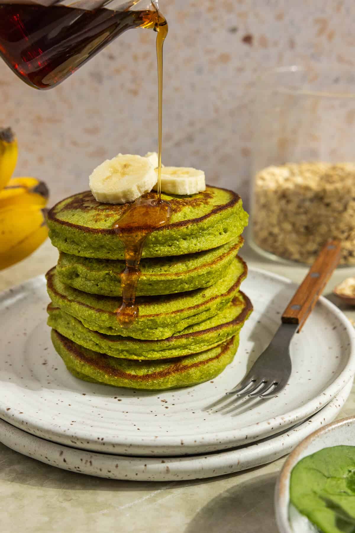 A plate with a stack of banana spinach pancakes topped with bananas and with maple syrup being drizzled over the top.