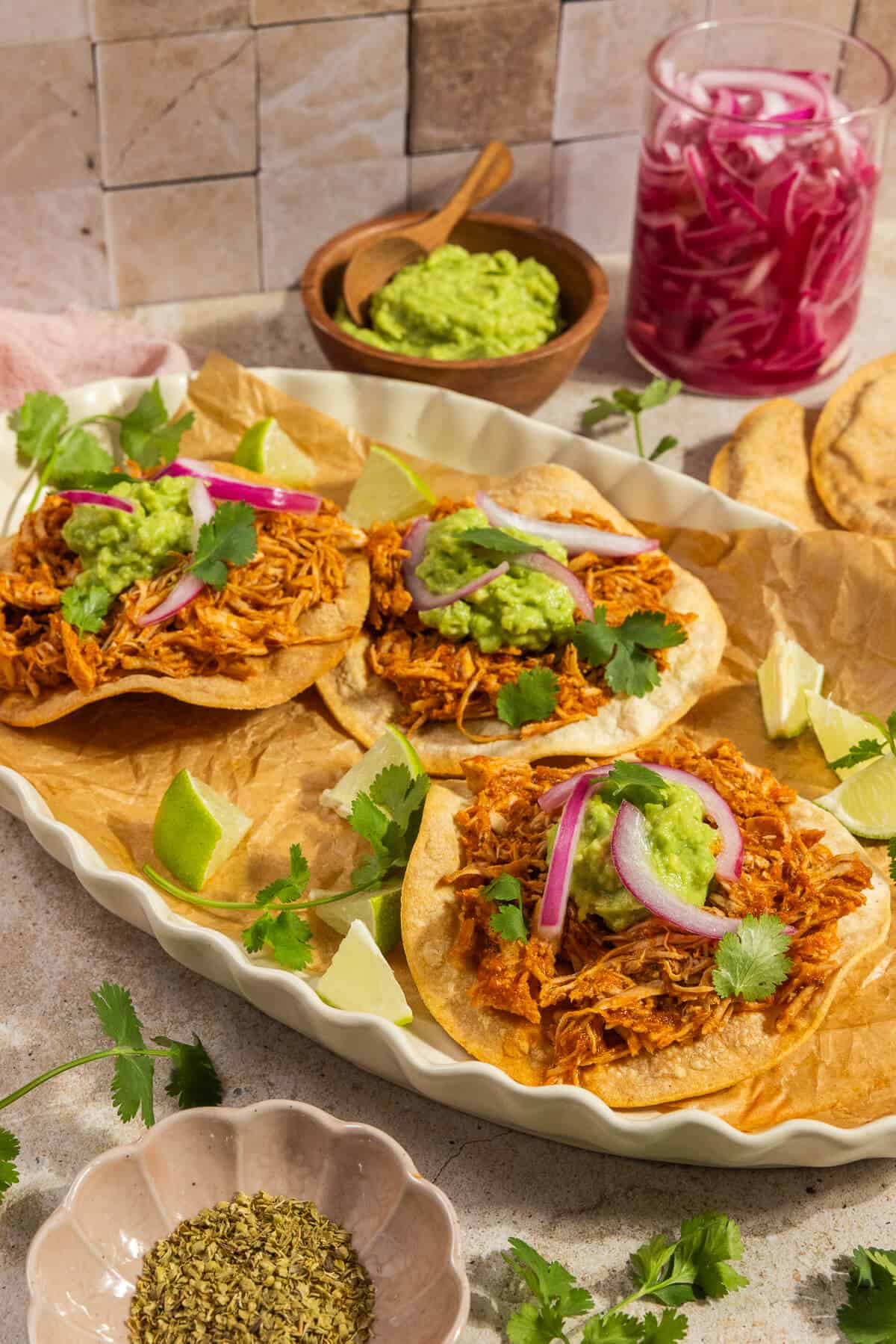 A platter with three chicken tinga tostadas topped with guacamole, pickled onions, and cilantro.