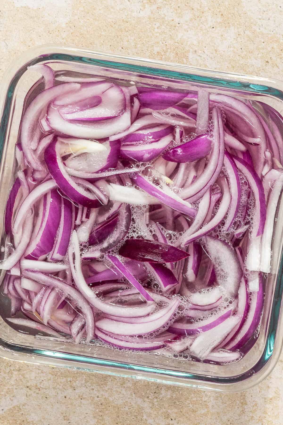 A Tupperware container with sliced red onions in a pickling solution.