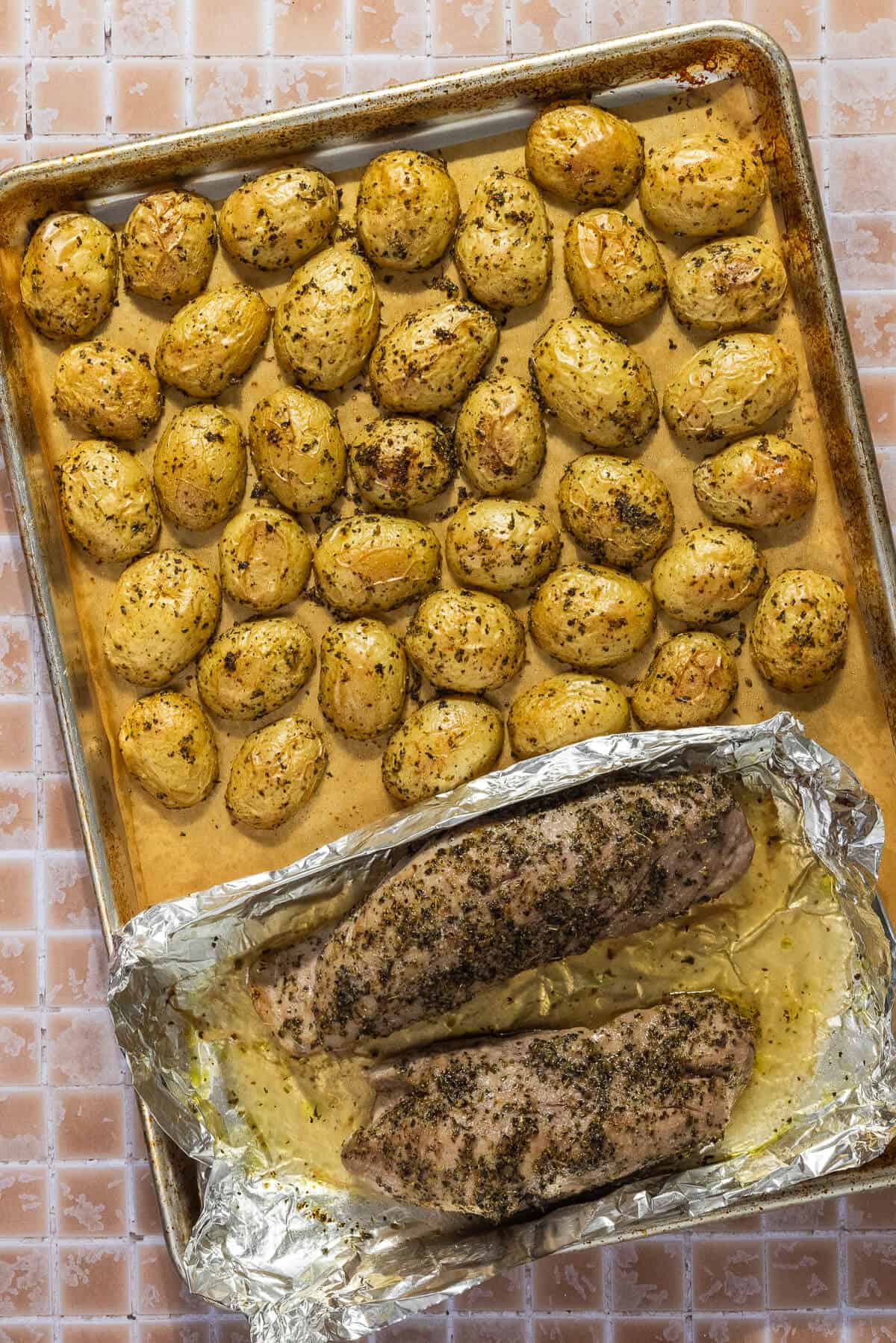 A parchment lined sheet pan with roasted baby potatoes and two pieces of a pork tenderloin encompassed with foil to prevent leakage.