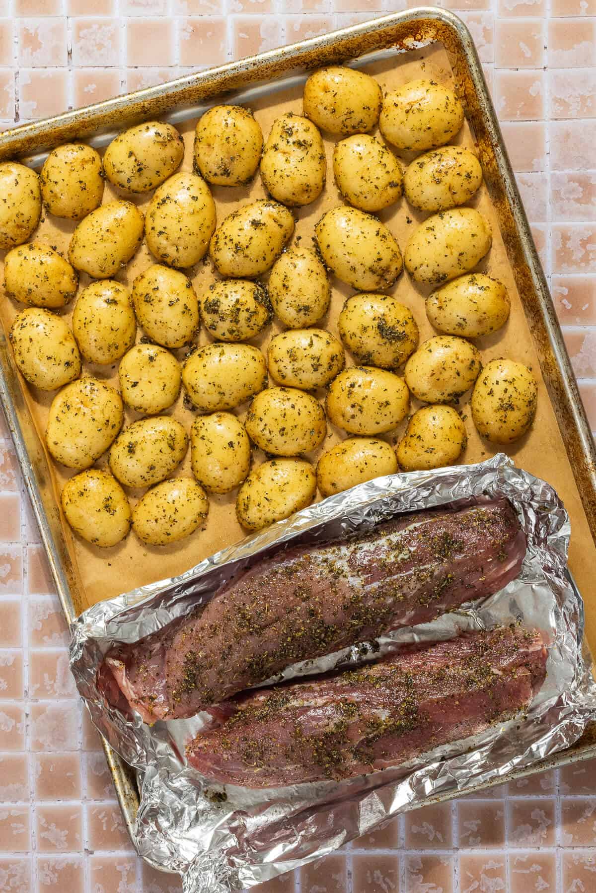 A parchment lined baking sheet with seasoned baby potatoes and pork tenderloin, with the pork tenderloin on top of foil to prevent leakage onto the potatoes.